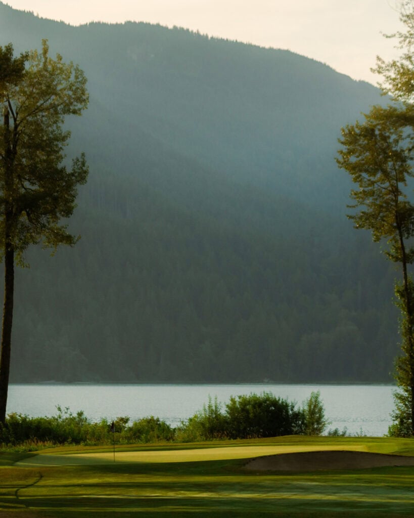 A serene golf green with a flag is in the foreground, bordered by trees and shrubs, with a calm lake and forested mountains in the background under a hazy sky—perfect for a peaceful day of golf amidst nature’s beauty.