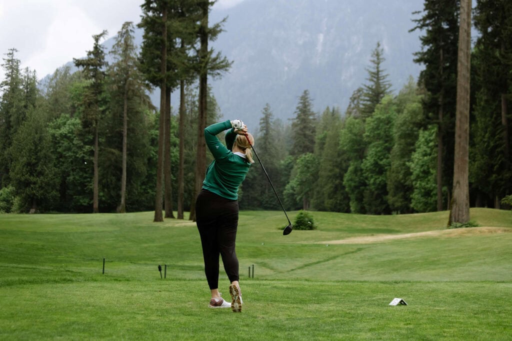 Person in green long-sleeve shirt and black pants swings a golf club on a green golf course surrounded by tall trees and mountains.