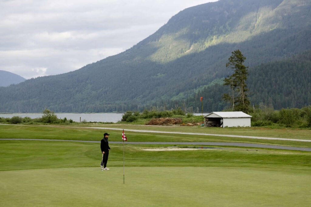 A person stands near a golf flag on a green field with mountains, trees, a small building, and a lake in the background under a cloudy sky.