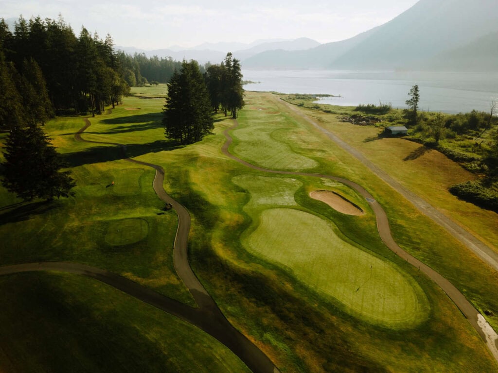 Aerial view of a golf course by a lake, showing fairways, greens, sand bunkers, and tree-lined areas with mountains in the background.