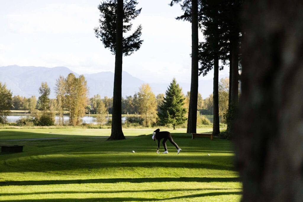 A person bends down on a grassy golf course surrounded by tall trees, with a lake and mountains visible in the background.