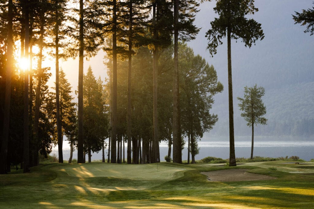 Sunlight filters through tall trees onto a golf course with a sand trap, with a lake and forested hills in the background.