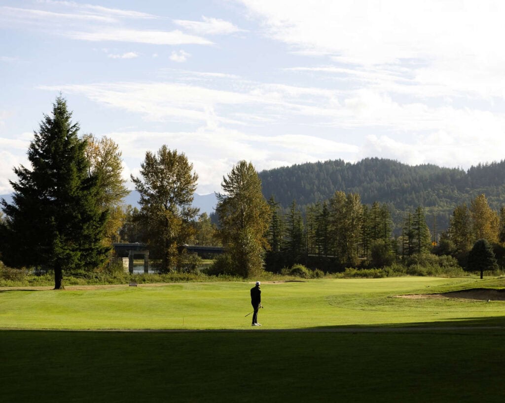 A person walks alone on a golf course surrounded by trees and hills under a partly cloudy sky.