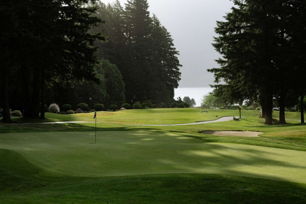 A golf course with a green, a flagstick, sand bunkers, and tall trees on both sides under a cloudy sky.