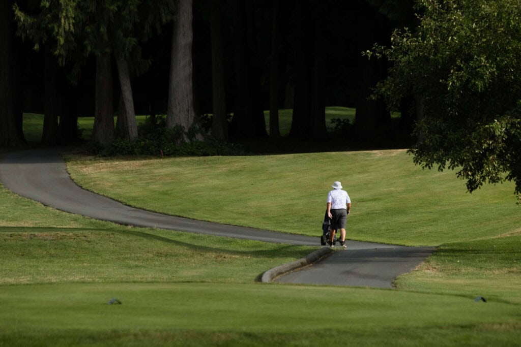 A person wearing a white hat walks alone on a golf course path, pulling a golf bag in the sunlight with trees in the background.