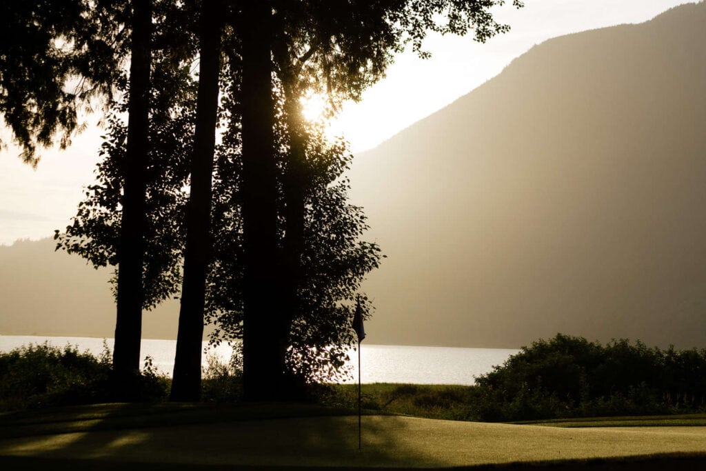 Sunlight filters through tall trees beside a golf green, with a flagstick visible and a mountain and lake in the background.