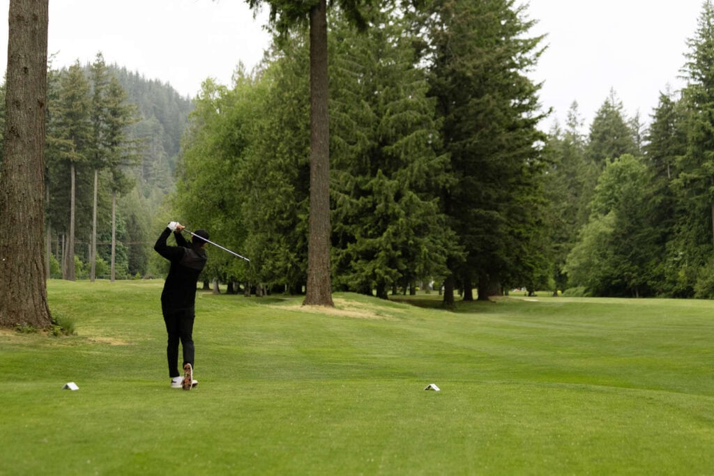 A person in dark clothes swings a golf club on a green golf course surrounded by tall trees and forested hills.